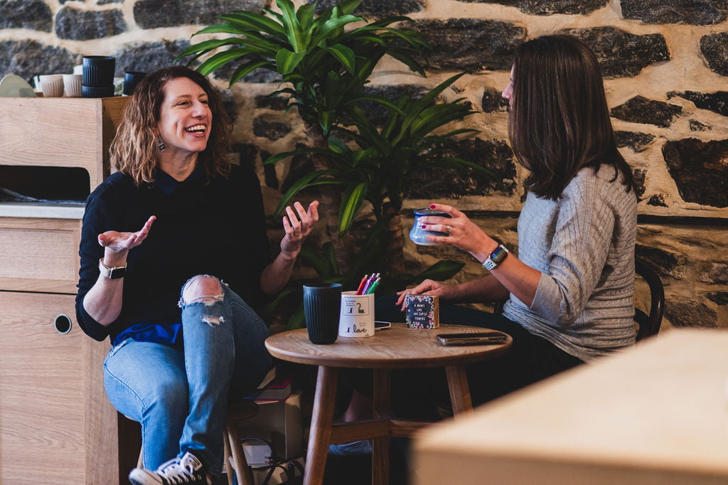 Jess Gill and Elisa Watson of Sunny Side Creative sitting and talking at a small table with coffee mugs