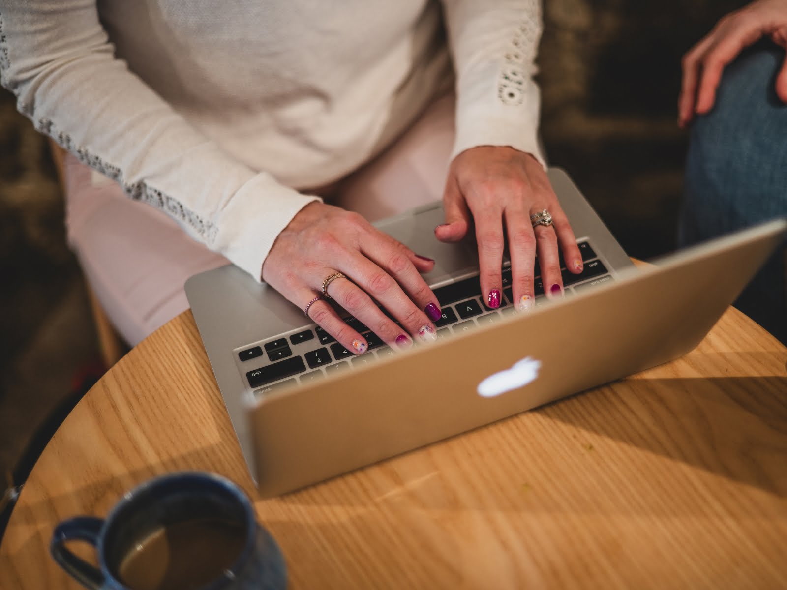 Overhead view of a woman's torso and hands at a table with a Macbook and coffee mug on the table