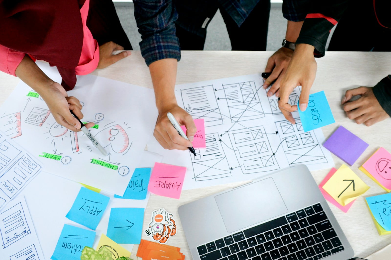 An overhead shot of people writing down ideas and strategizing with post-its, wireframes, and a computer.