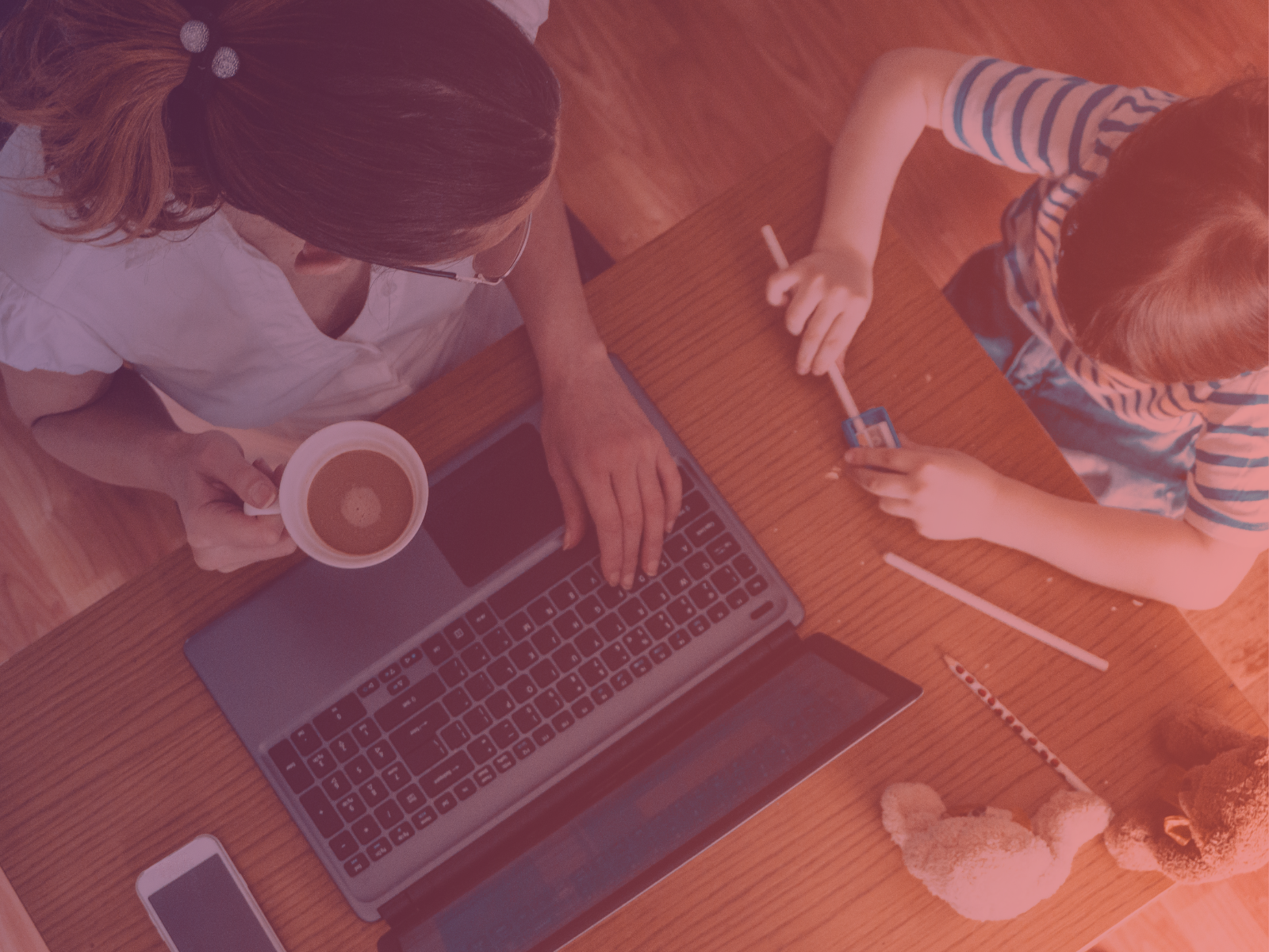A woman works on a laptop, holding coffee, while a child beside her plays with pencils and a sharpener on a wooden table. Stuffies are nearby, depicting balancing motherhood and work.