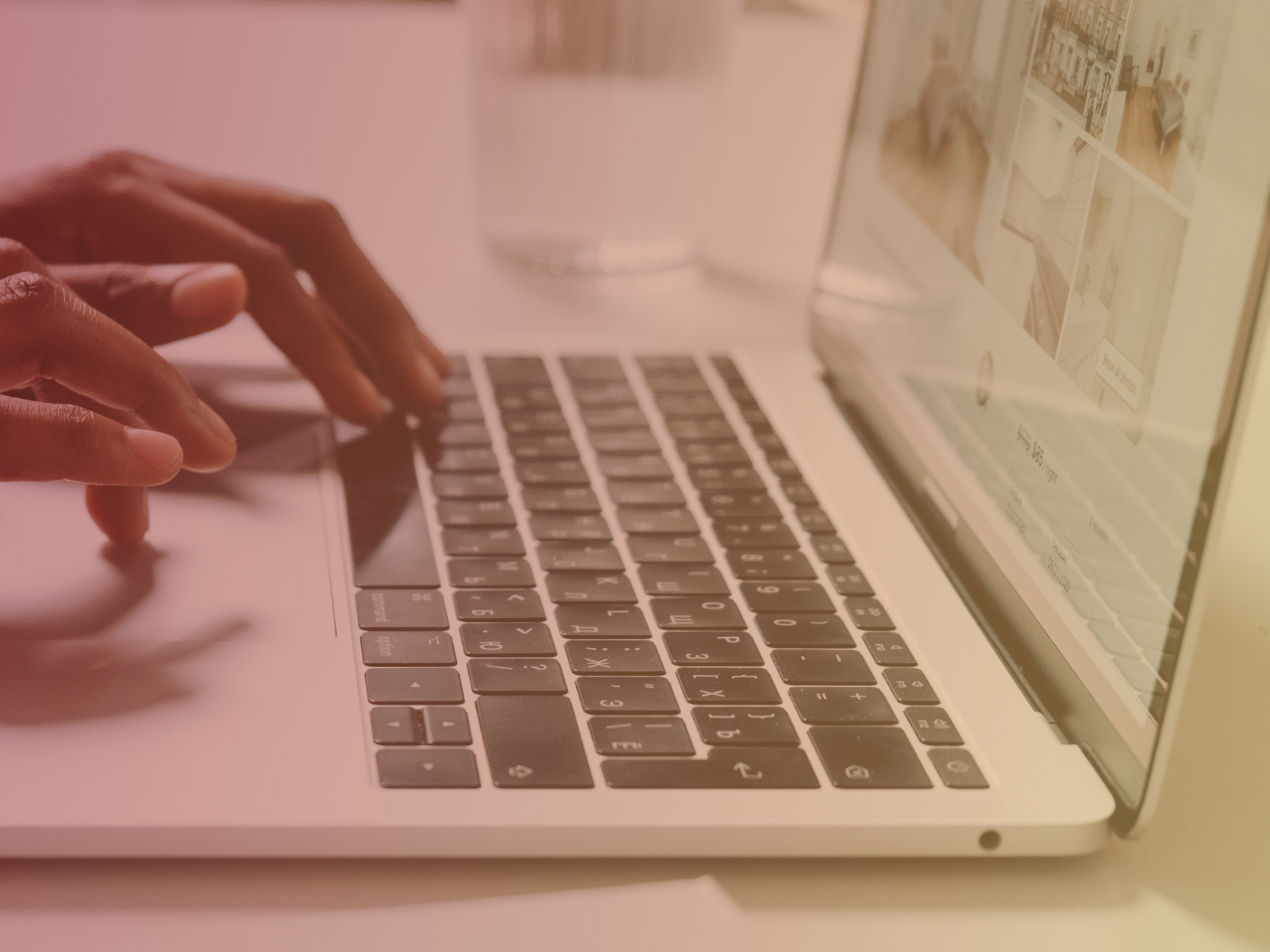 A person types on a laptop keyboard with a glass of water and a blurred website visible on the screen in the background.